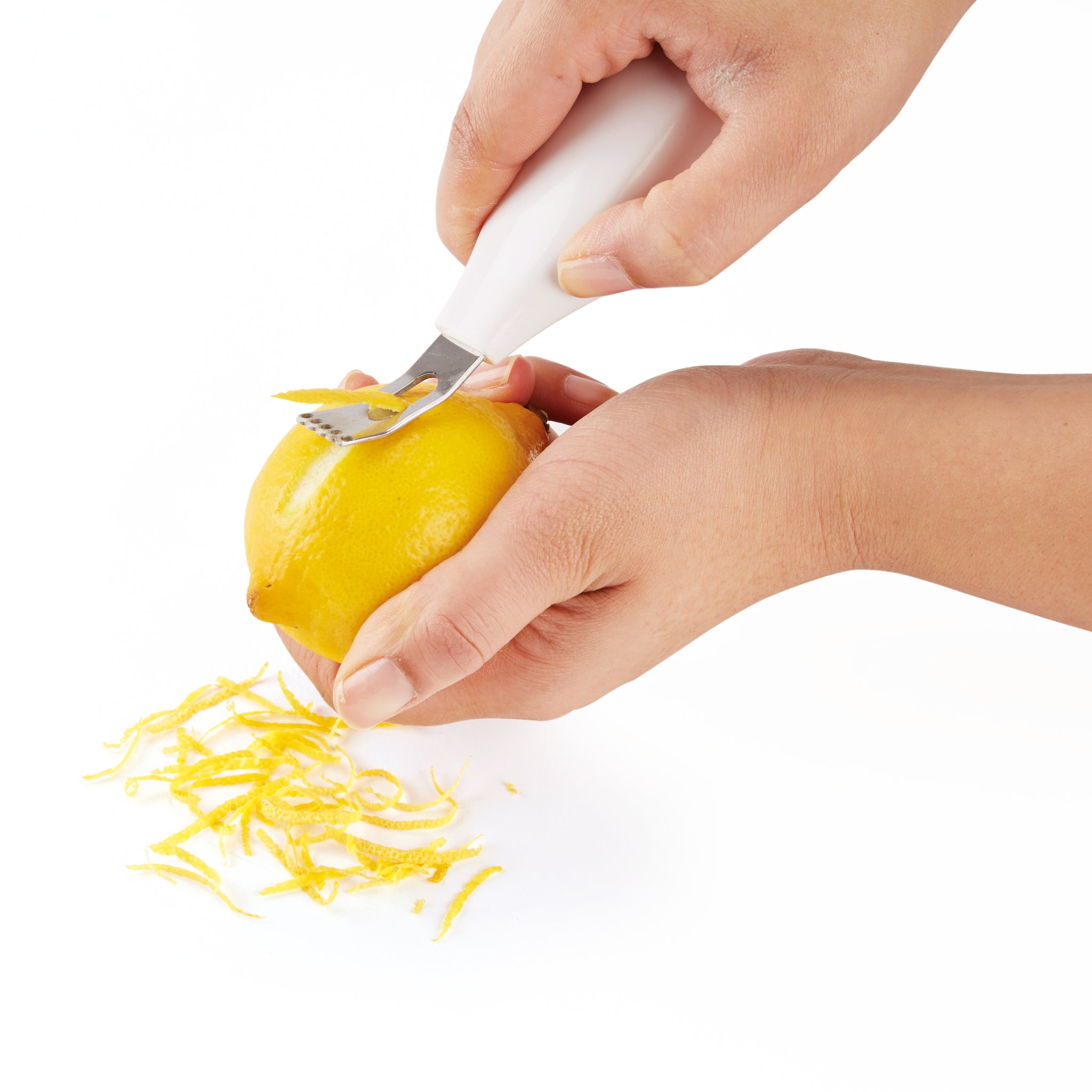 A person uses a white-handled zesting tool to peel thin strips of zest from a yellow lemon, with zest shavings gathered below on a white surface.