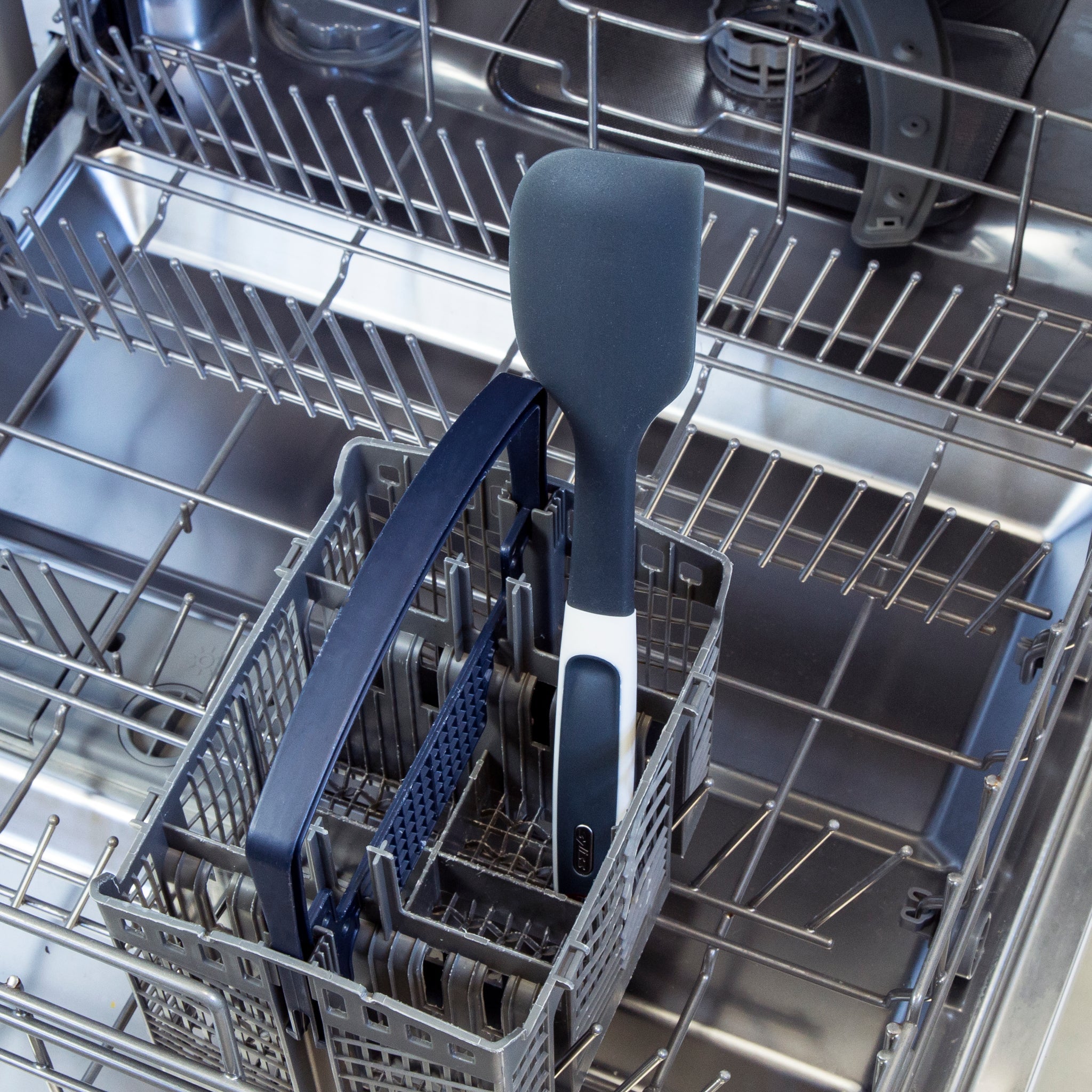 A blue and white spatula placed in the utensil basket of an open dishwasher with empty racks above and around it.