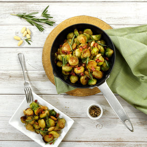 Brussels sprouts with bacon and herbs are perfectly cooked in the Zyliss Ultimate Pro Non-Stick Frying Pan With Pouring Lip, shown on a wooden board beside a served dish, black pepper, pine nuts, and rosemary atop a white wooden table. #20cm/8"