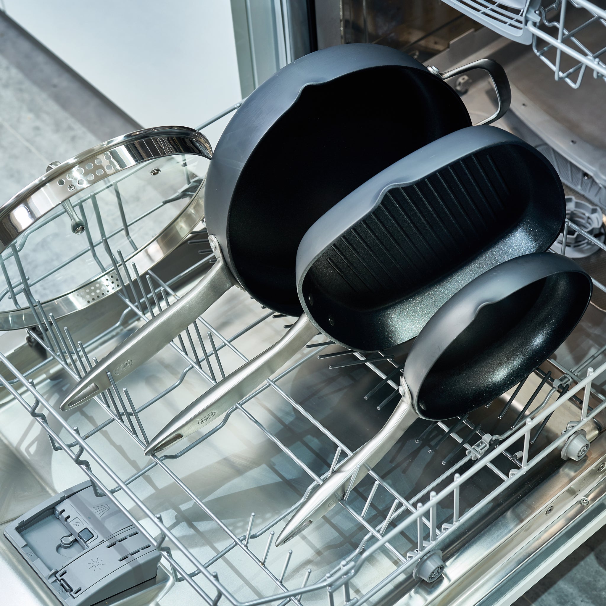 Three nonstick black pans and two glass lids are arranged in the bottom rack of an open dishwasher, ready to be cleaned.