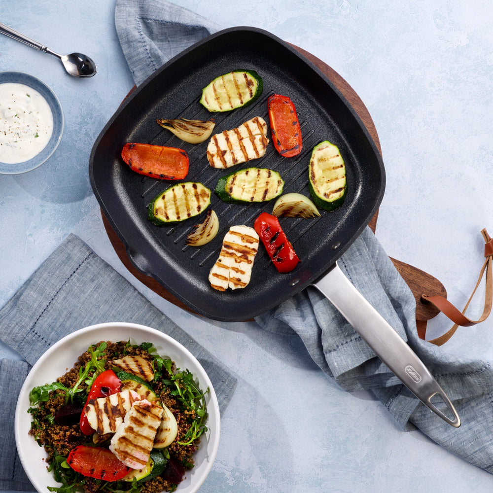 The Zyliss Ultimate Pro Non-Stick Square Grill Pan With Pouring Lip, black with raised ridges and a long silver handle, made from hard-anodized aluminium, is set against a plain light background. 