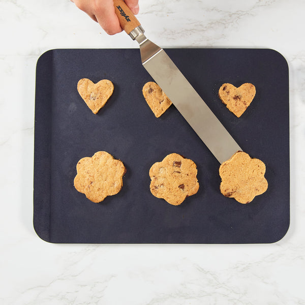 A hand uses the Zyliss Beech Wood Angled Palette Knife to lift a flower-shaped chocolate chip cookie from a baking sheet holding three flower and three heart cookies on a white countertop. 