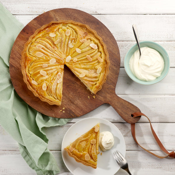 A pear tart with almond slices, baked in the Zyliss Durable Non-Stick Tart Pan, sits on a wooden board with one slice missing. Nearby are a plated slice with whipped cream, extra whipped cream, and a green cloth on a white table. 