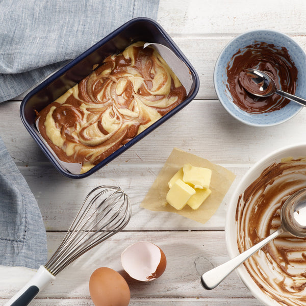 A Zyliss Durable Non-Stick Loaf Pan filled with swirled marble cake batter rests on a white wooden table, surrounded by chocolate, butter on parchment, a whisk, two eggs (one cracked), and a bowl with leftover batter. #1L/1lb