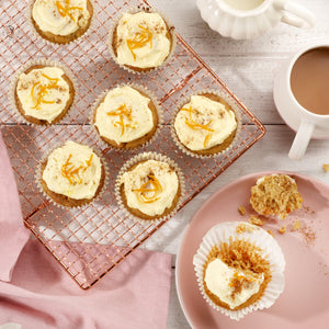 Cream-frosted cupcakes with orange zest cool on a Zyliss Durable Non-Stick 12 Hole Muffin Pan, beside a cup of tea, a milk jug, and a crumb-dusted pink plate on a light wood surface. 