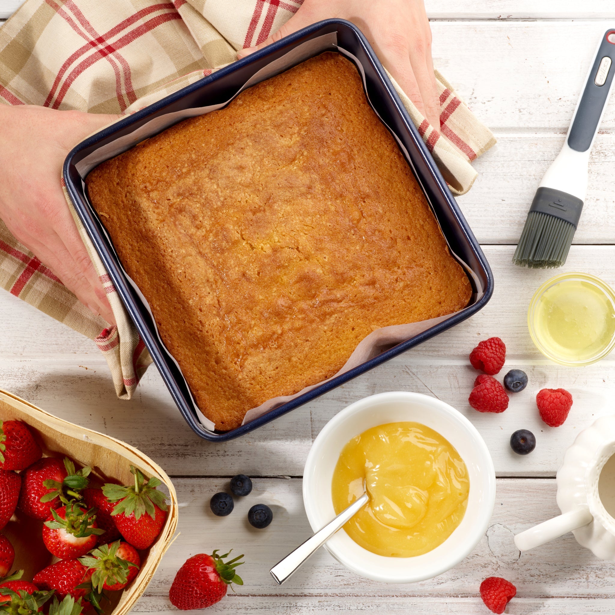 A person holds a square sponge cake in a pan on a wooden table with a bowl of lemon curd, a basket of strawberries, loose raspberries and blueberries, a pastry brush, and a checked cloth nearby.