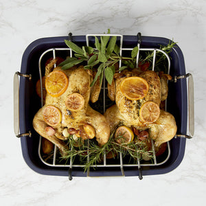 Two roasted chickens sit in the Zyliss Durable Non-Stick Roasting Pan, garnished with lemon slices and fresh herbs on a metal rack above vegetables. Dishwasher safe for easy cleanup. Marble countertop background. 