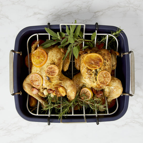 Two roasted chickens sit in the Zyliss Durable Non-Stick Roasting Pan, garnished with lemon slices and fresh herbs on a metal rack above vegetables. Dishwasher safe for easy cleanup. Marble countertop background. 
