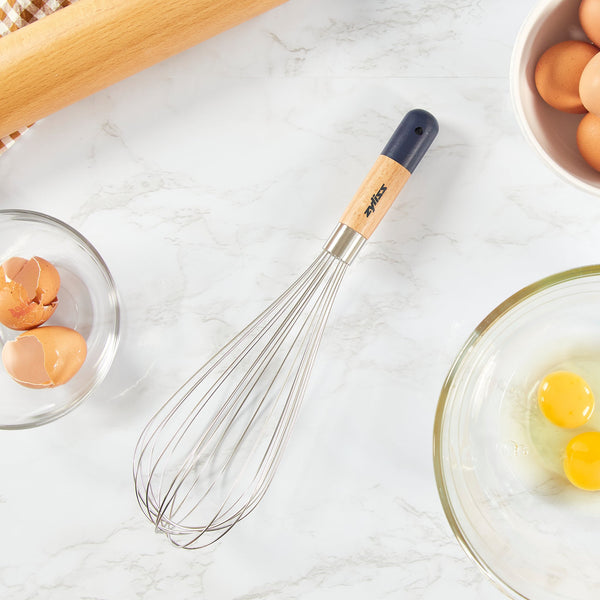 The Zyliss Beech Wood Balloon Whisk, featuring stainless steel wires and a beech wood handle, rests on a marble countertop beside a rolling pin, whole eggs, cracked eggs, and a glass bowl with two cracked eggs. 
