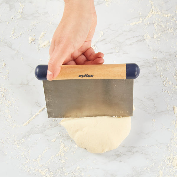A hand uses the Zyliss Beech Wood Bench Scraper to divide dough on a floured surface, demonstrating this essential baking tool in use. 