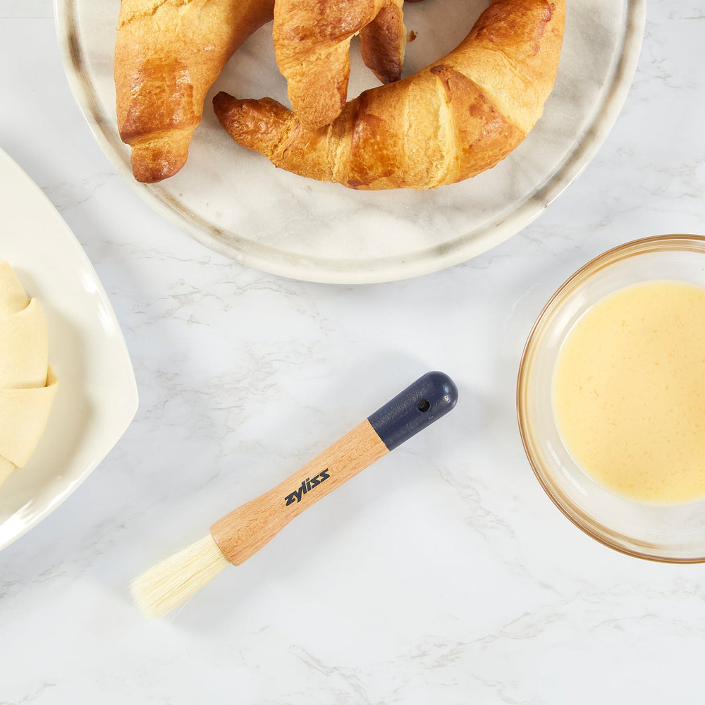 The Zyliss Beech Wood Pastry Brush features natural bristles, a beech wood handle with a printed logo, dark blue top, and hanging hole, shown on a plain white background. 