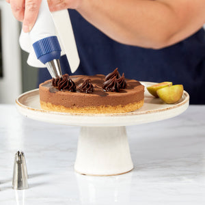 Using the Zyliss Reusable Piping Bag Kit, someone pipes chocolate cream onto a chocolate tart. The tart is displayed on a white cake stand and garnished with a halved yellow plum. 