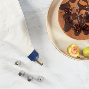 The Zyliss Reusable Piping Bag Kit, with a metal tip and four stainless steel nozzles, rests on marble beside a sliced fig and a chocolate cake adorned with frosting on a beige plate. 