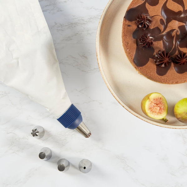 The Zyliss Reusable Piping Bag Kit, with a metal tip and four stainless steel nozzles, rests on marble beside a sliced fig and a chocolate cake adorned with frosting on a beige plate. 