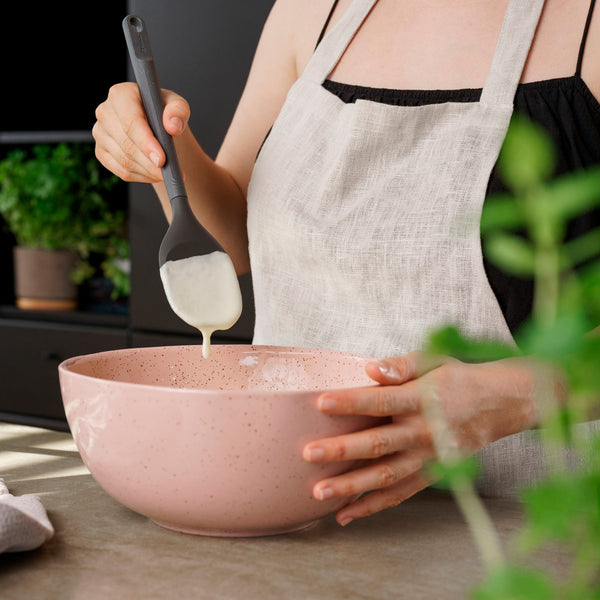 Wearing a light apron, a person stirs a creamy mixture in a pink bowl using the Zyliss Spoontula, noted for its ergonomic handle, with green plants in the background. #Large