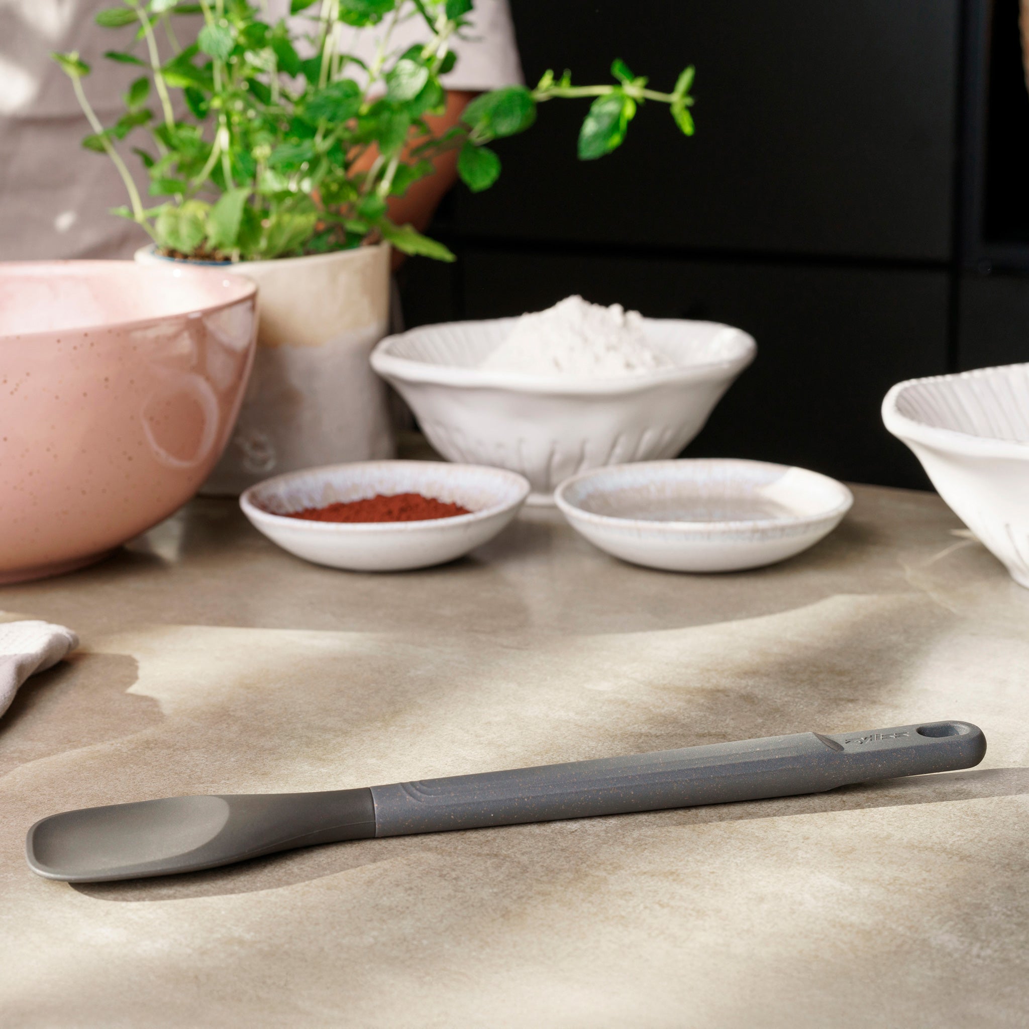 A Zyliss Spoontula with a gray silicone head and ergonomic handle rests on a kitchen counter, surrounded by bowls of flour, cocoa powder, other ingredients, a pink mixing bowl, and a potted herb plant in the background. #Medium