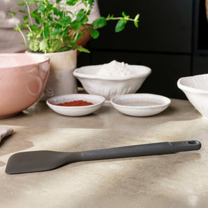 A black, heat-resistant Zyliss Spatula with a silicone head is on the kitchen counter near bowls of flour and cocoa powder, with a pink mixing bowl and potted herb in the background. #Large