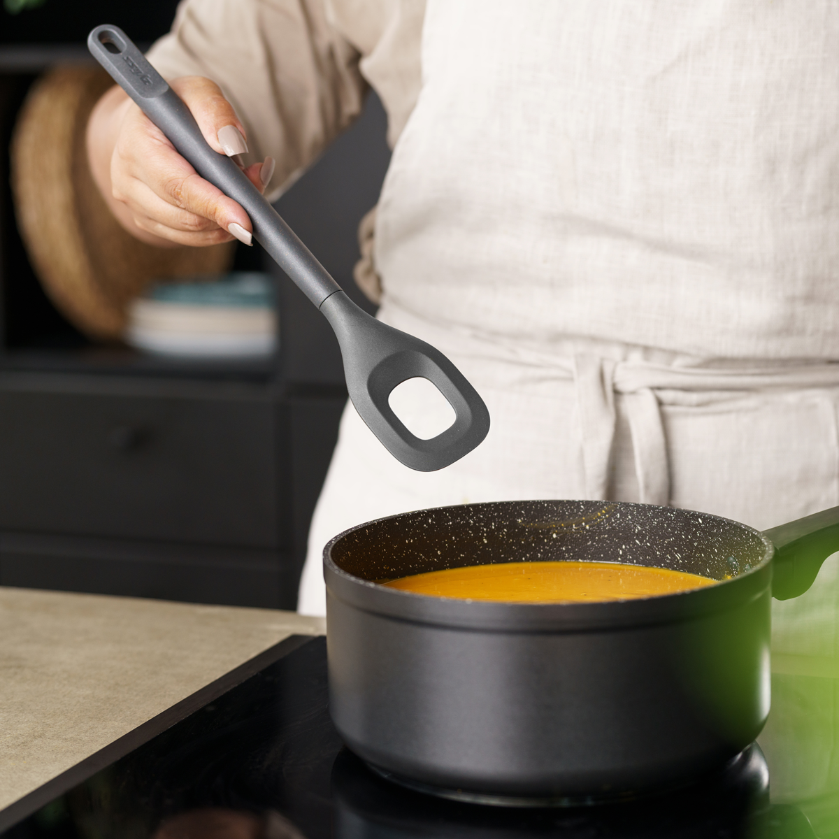A person wearing a light apron is stirring a pot of orange soup on a stovetop with a black cooking utensil. The scene appears to be in a modern kitchen.