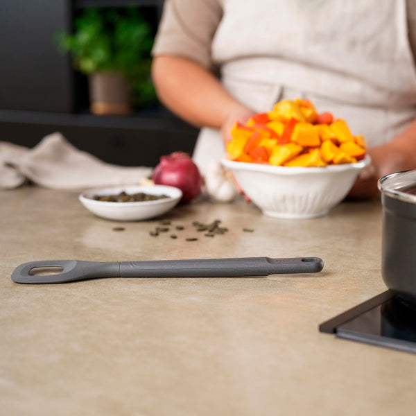 A gray Zyliss Square Mixing Spoon, heat-resistant and dishwasher safe for easy cleanup, rests on the kitchen counter. Nearby are chopped orange vegetables in a bowl, an onion, seeds, and a pot on the stove. 