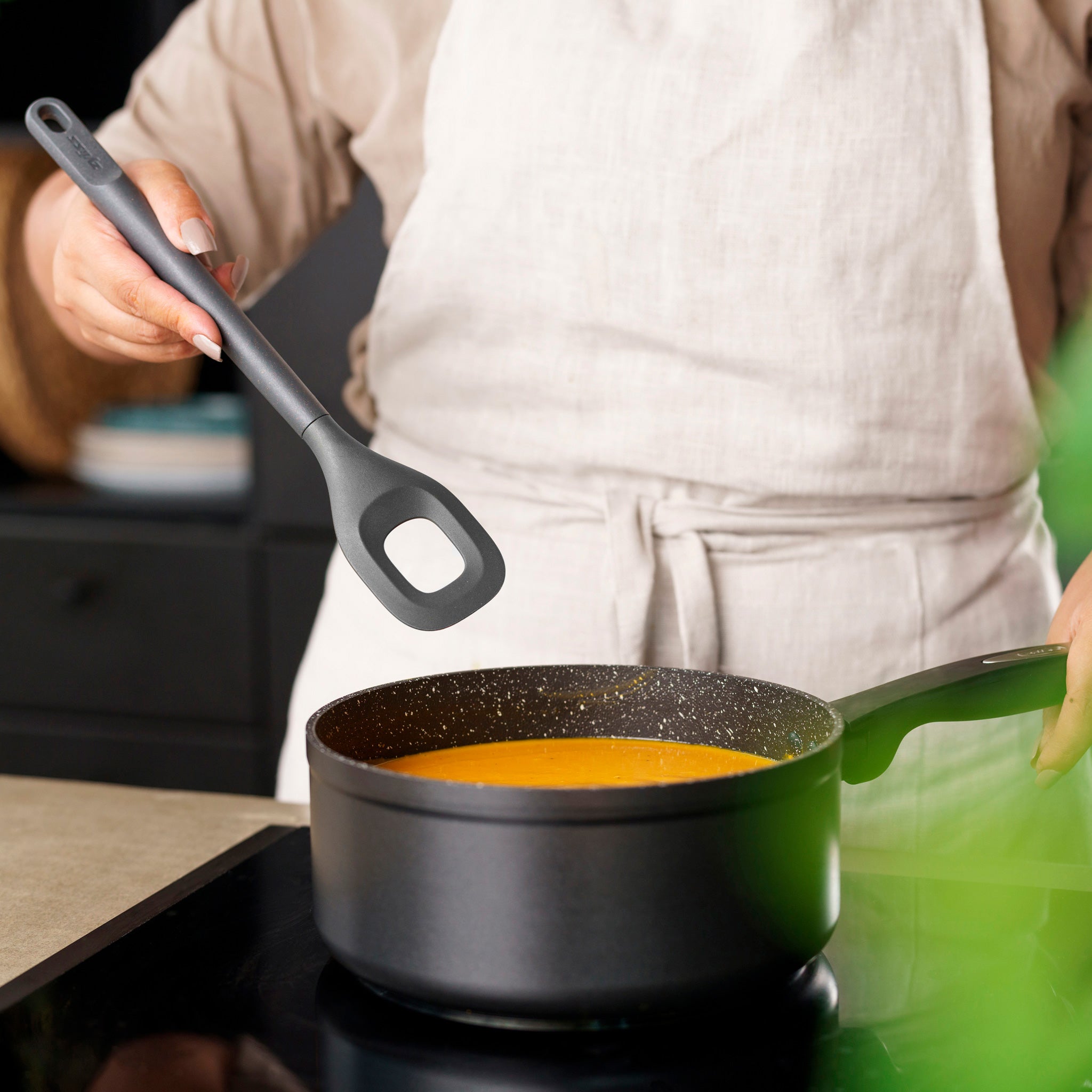 A person in a light-colored apron holds a Zyliss Square Mixing Spoon above a saucepan of orange soup on the stove, with only their torso and hands visible.