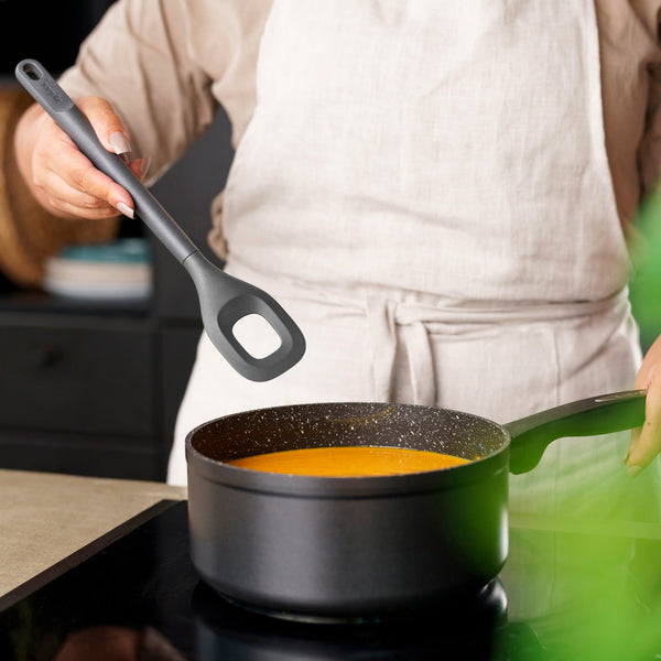 A person in a light-colored apron holds a Zyliss Square Mixing Spoon above a saucepan of orange soup on the stove, with only their torso and hands visible. 