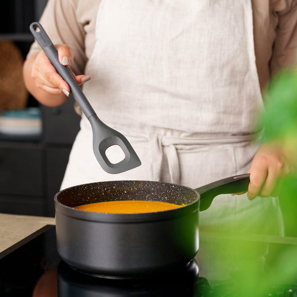 A person stirs orange soup in a black non-stick saucepan on the stove, using a Zyliss Angled Mixing Spoon with an ergonomic handle and a square hole in the center, while wearing a light apron. 