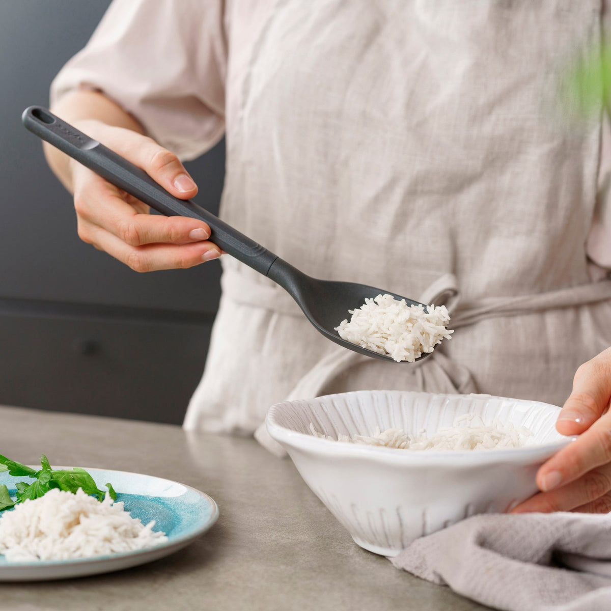 Wearing a light shirt, a person uses a durable, heat-resistant Zyliss Spoon to serve cooked white rice into a white bowl on the kitchen counter, next to a small plate of herbs. #Medium