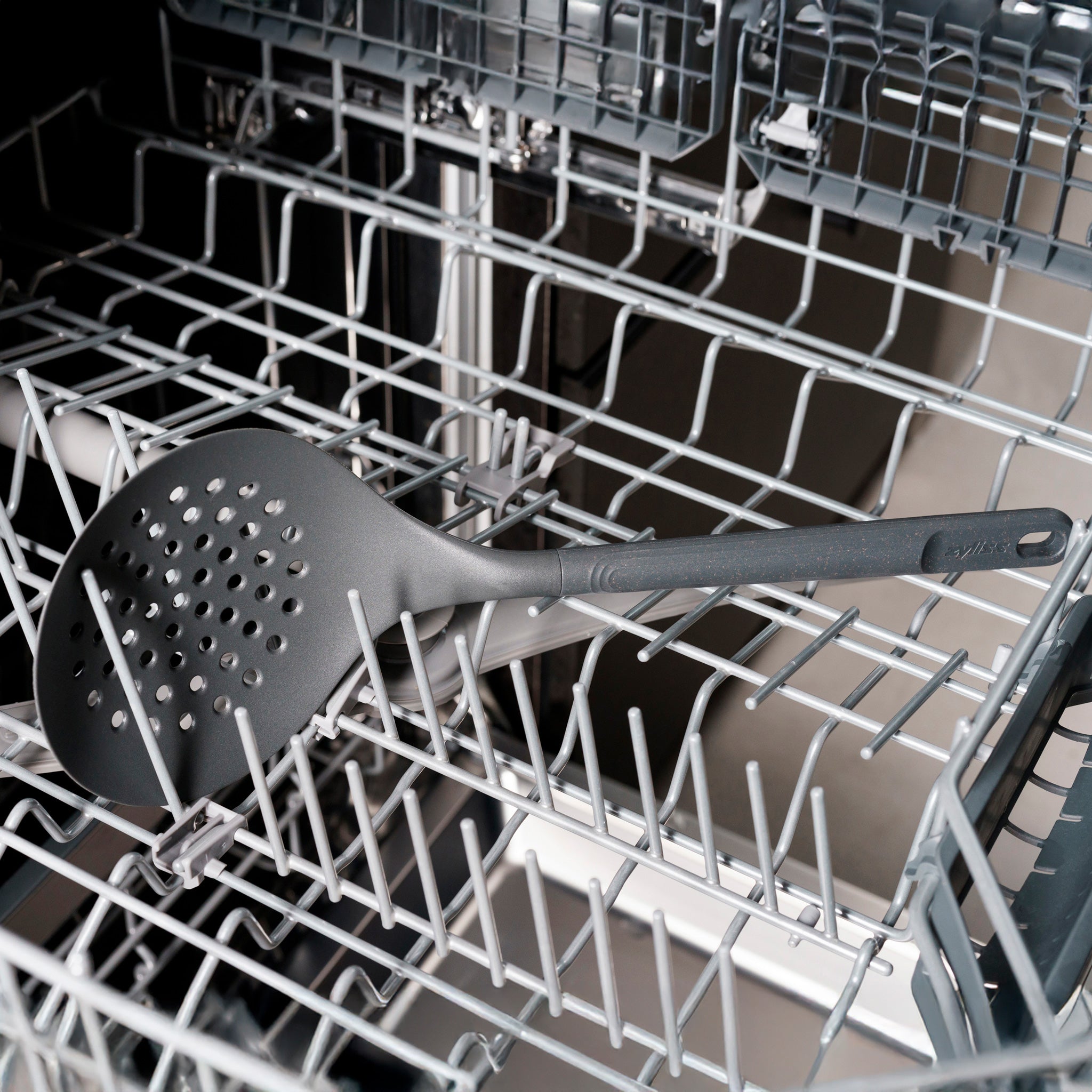 A large black slotted spoon rests diagonally across the top rack of an open dishwasher, surrounded by metal prongs designed to hold dishes.