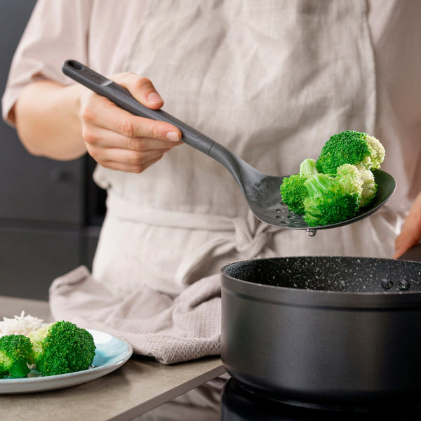 Wearing an apron, a person uses a Zyliss Skimmer to drain and serve steamed broccoli from a black pot, with extra broccoli on a plate nearby. 