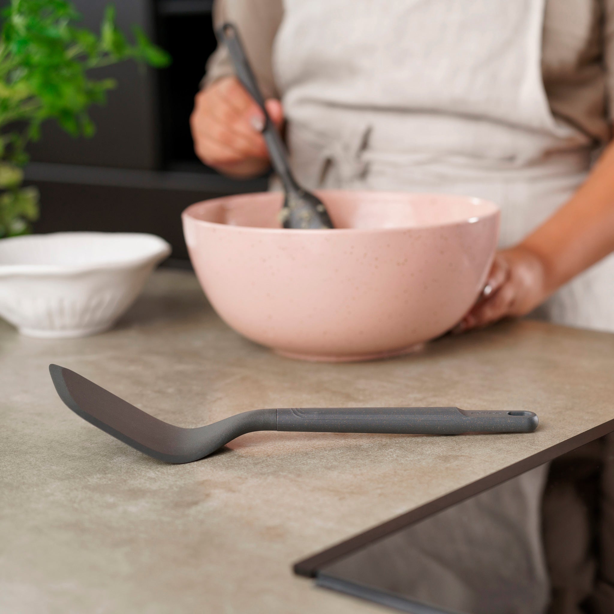 Wearing an apron, a person mixes ingredients in a pink bowl with a spatula as the Zyliss Angled Turner rests on the kitchen counter. A white bowl and green plant are also visible.