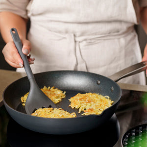 Wearing a beige apron, a person cooks golden-brown potato pancakes in a black frying pan on the stove, using the Zyliss Slotted Turner with an ergonomic handle to flip one of the pancakes. 