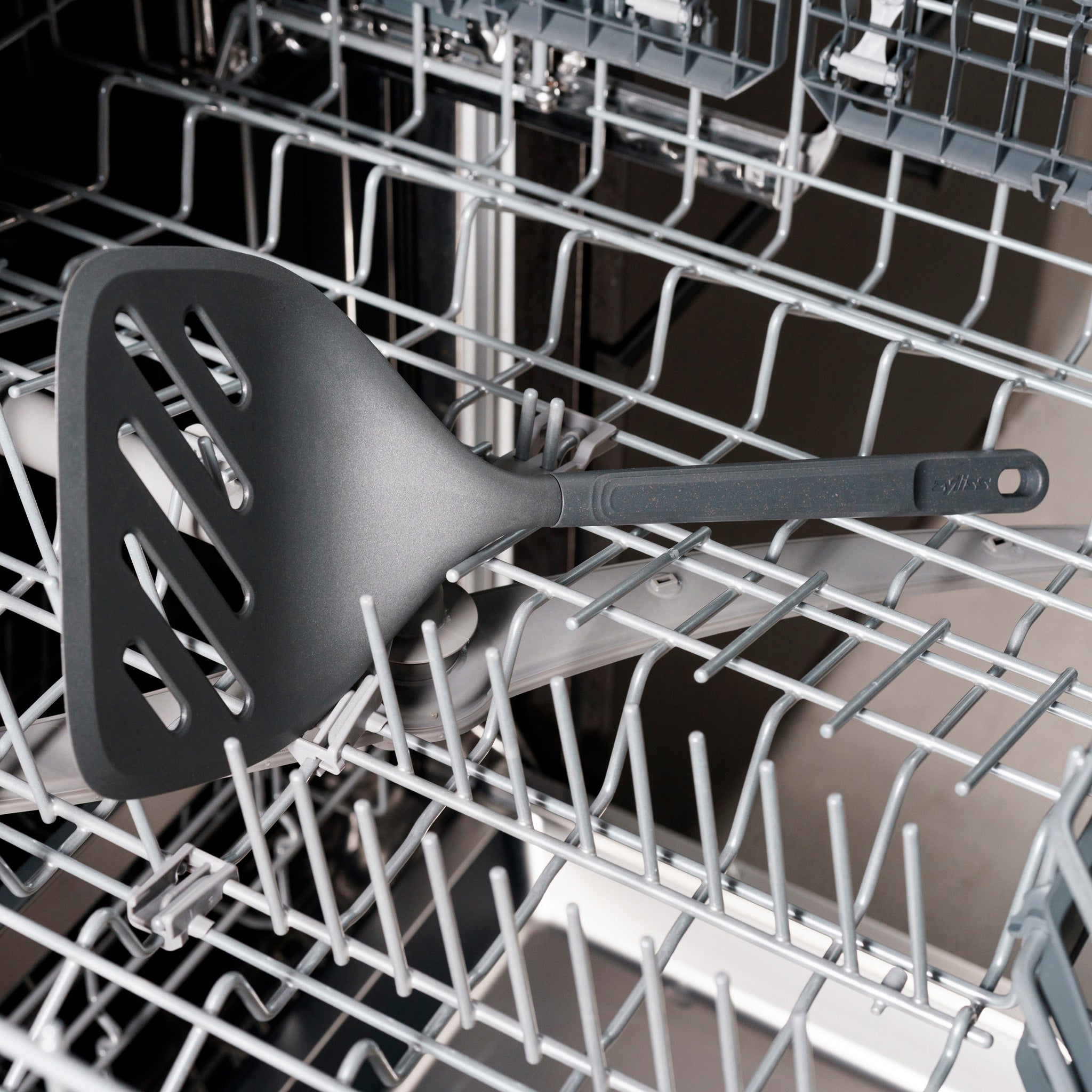 A black slotted spatula is placed on the top rack of an open dishwasher, surrounded by metal racks and no other dishes.