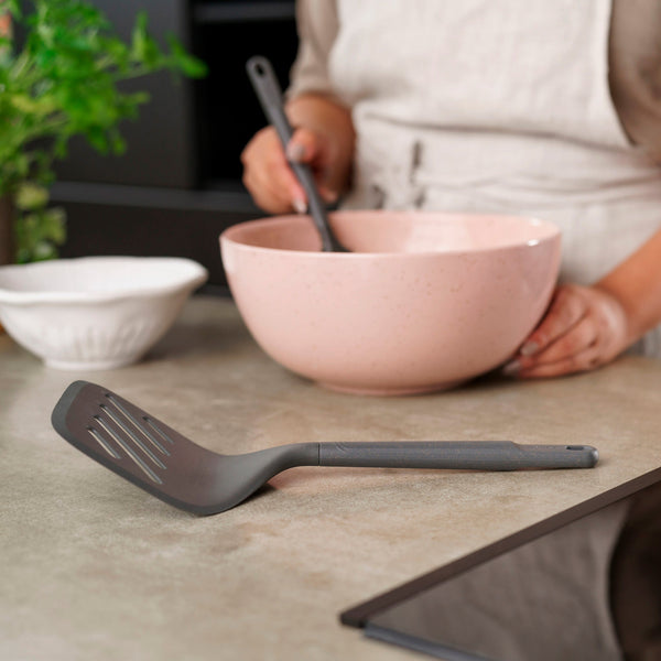 Wearing an apron, a person stirs a pink mixing bowl on the kitchen counter as the Zyliss Turner XL, a heat-resistant turner with an ergonomic handle, rests in the foreground. Also visible are a white bowl and a green plant. 