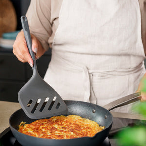 Wearing a light apron, a person uses the Zyliss Turner XL to lift or flip a golden-brown hash brown in a nonstick frying pan on the stove. 