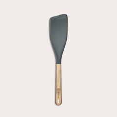 The Zyliss Oak Wood Easy Lift Turner has a dark gray silicone head, an oak wood handle, and a hanging hole at the end. It is shown on a plain light background.