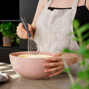 Wearing a linen apron, a person mixes batter in a large pink bowl with green herbs nearby using the Zyliss Balloon Whisk, which is safe for non-stick cookware. #Medium