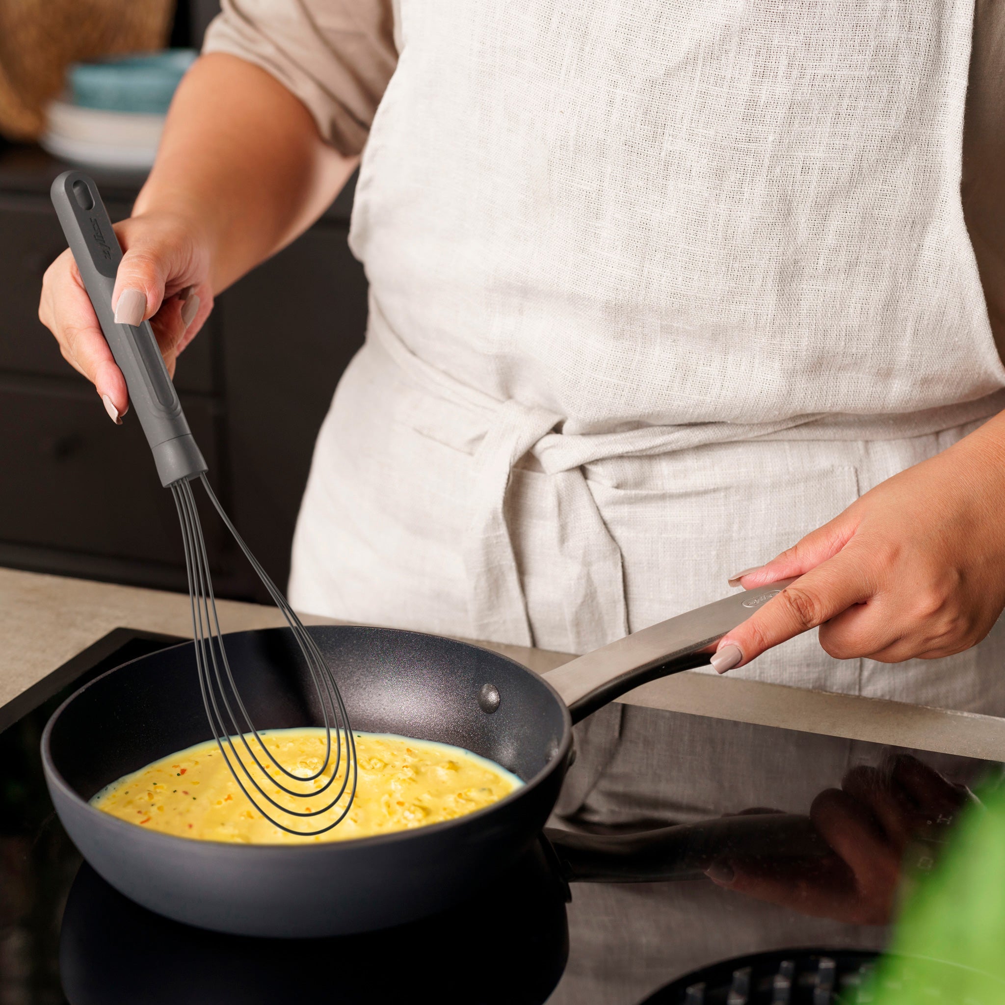Wearing a light-colored apron, someone uses the Zyliss Flat Whisk Silicone to stir eggs in a nonstick frying pan on the stovetop.