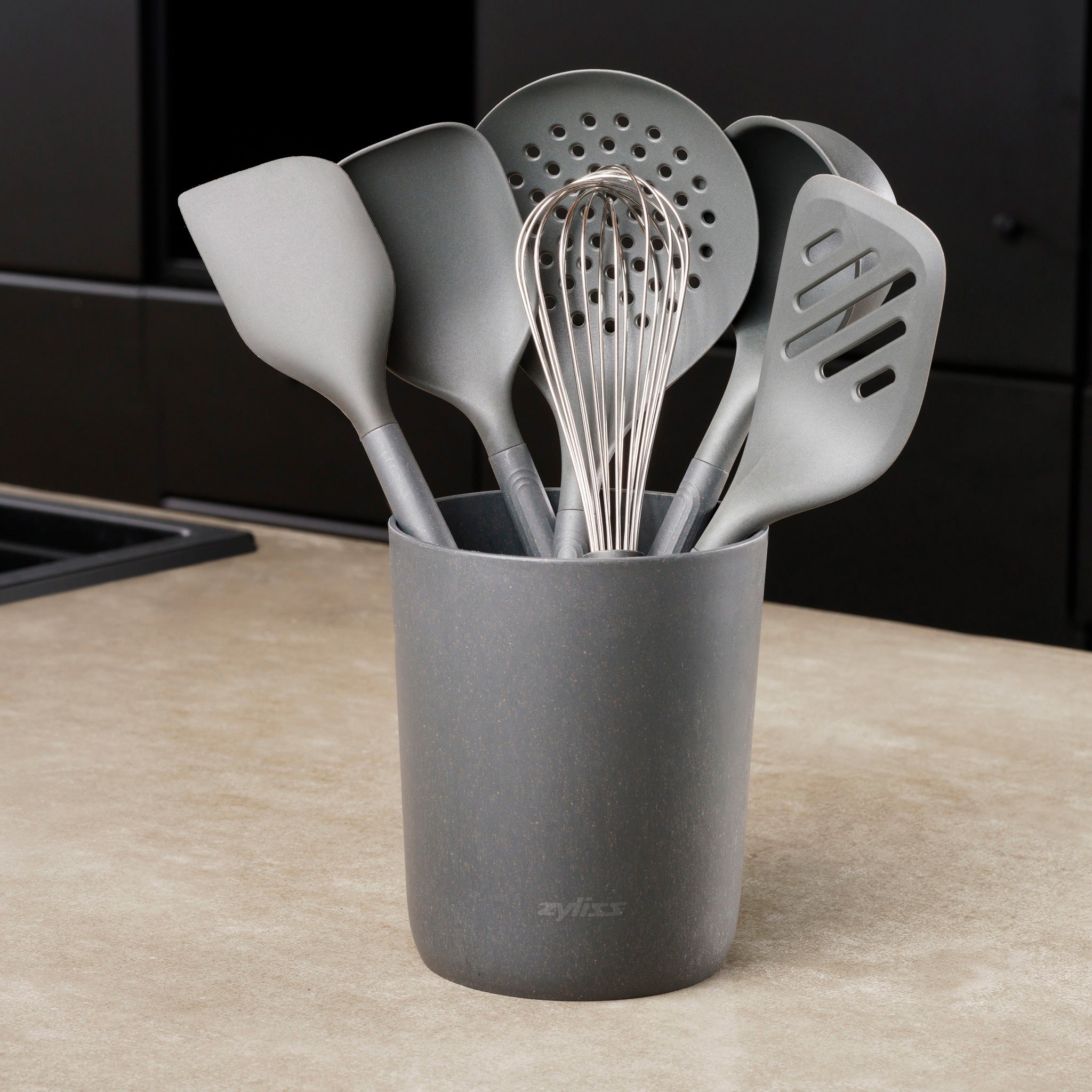 A gray Zyliss 7-Piece Utensil Set with ergonomic handles rests on the counter, holding spatulas, a slotted spoon, whisk, and ladle—all in heat-resistant gray PBT.