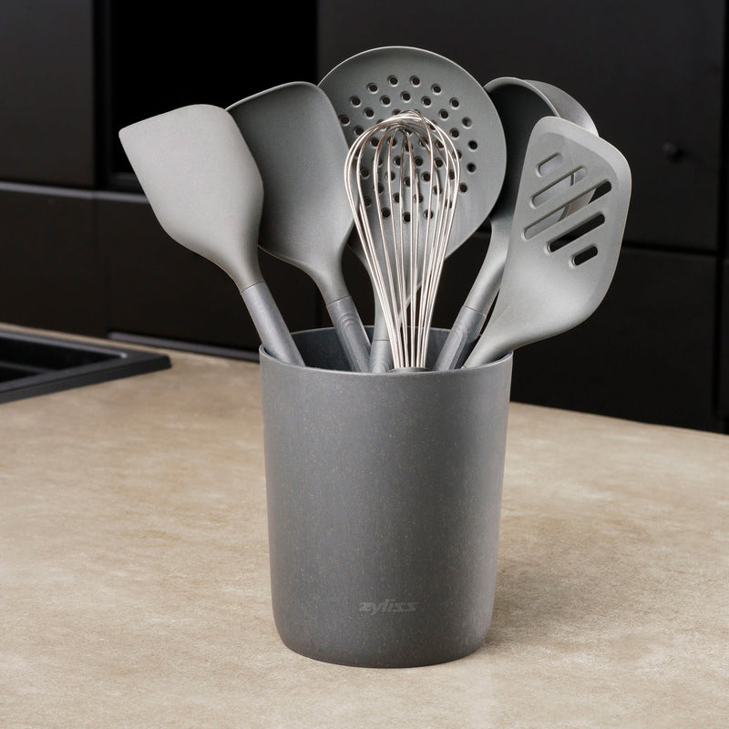 A gray Zyliss 7-Piece Utensil Set with ergonomic handles rests on the counter, holding spatulas, a slotted spoon, whisk, and ladle—all in heat-resistant gray PBT.