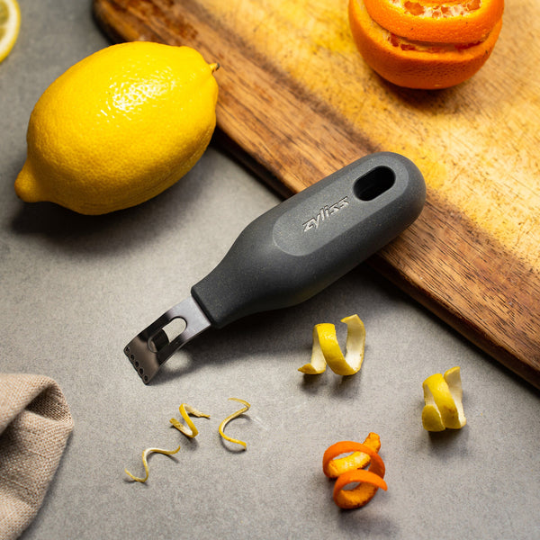 The Zyliss 2-In-1 Zester with a black handle rests on a gray surface alongside lemon and orange peels, a whole lemon, a partially zested orange, and a wooden cutting board. 
