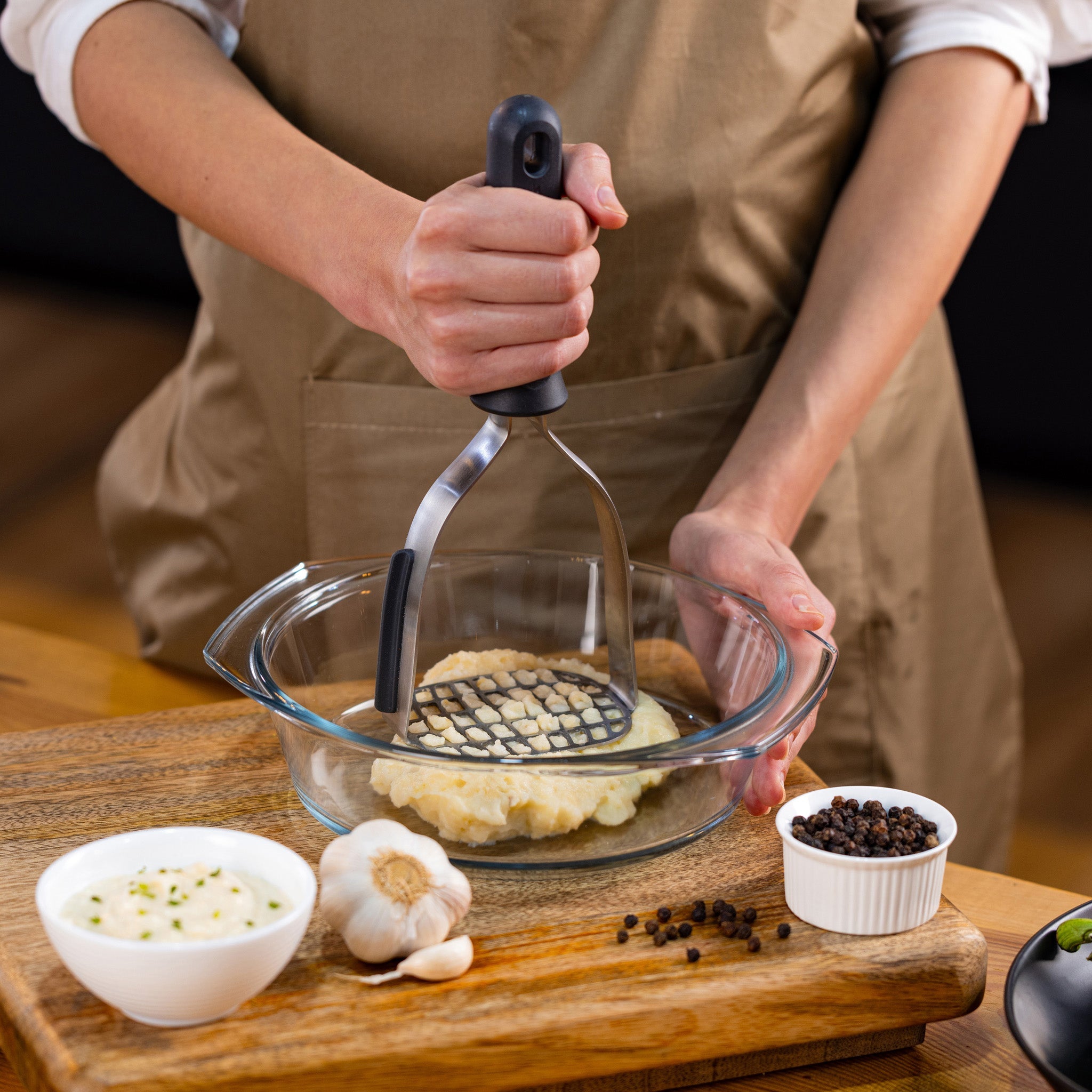 A person mashing potatoes in a glass bowl with a potato masher on a wooden table, surrounded by garlic, peppercorns, and a small bowl of creamy dip.