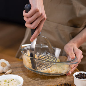 A person uses the Zyliss Potato Masher with a stainless steel head to mash cooked chickpeas in a glass bowl on a wooden surface, surrounded by garlic and small ingredient bowls. 