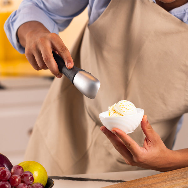 A person wearing a beige apron scoops vanilla ice cream into a white bowl, with a bowl of fresh fruit, including grapes and apples, visible on the counter nearby.