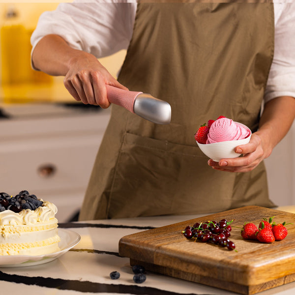 #pink A person in a brown apron uses the Zyliss Ice Cream Scoop to serve pink ice cream into a small bowl with strawberries next to a cutting board of berries and a cake on the kitchen counter.