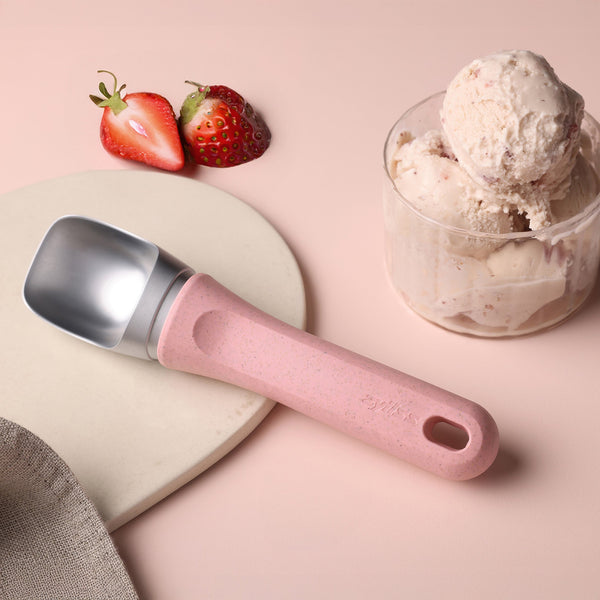 #pink A Zyliss Ice Cream Scoop rests beside a glass cup holding three scoops of strawberry ice cream and two strawberries, all placed on a light pink surface.