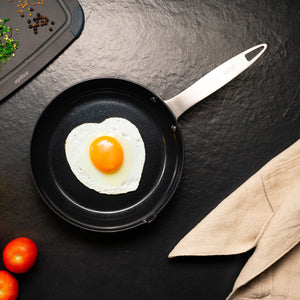 A heart-shaped fried egg in the Zyliss Ultimate Pro Ceramic Fry Pan with non-stick coating rests on a dark surface, surrounded by a beige kitchen towel, tomatoes, chopped herbs, and peppercorns. #20cm/8"
