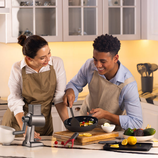 A woman and man in aprons smile and cook together in a modern kitchen. The man stirs food in the Zyliss Ultimate Pro Ceramic Fry Pan while the woman watches. Fresh ingredients and utensils are on the counter. #24cm/9½"