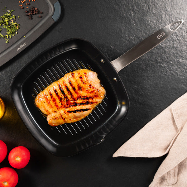 A grilled chicken breast with char lines cooks in a black grill pan on a dark countertop, surrounded by tomatoes, a beige cloth, and assorted cooking ingredients.