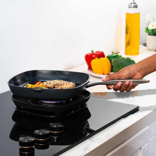 A hand holds the Zyliss Ultimate Pro Ceramic Square Grill Pan on a gas stove, cooking chicken and vegetables. On the counter are bell peppers, broccoli, a small plant, and oil in a clean, modern kitchen. 