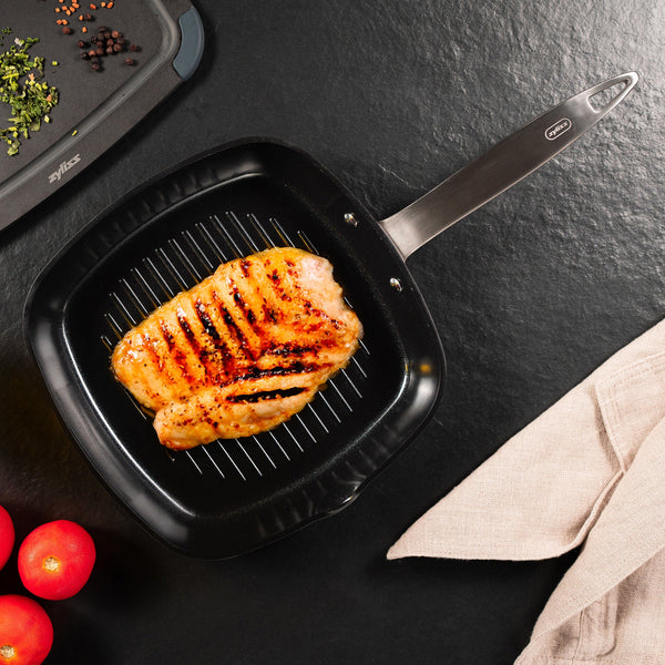 The Ultimate Pro Ceramic Square Grill Pan by Zyliss features a non-stick ceramic coating and stylish grill marks—ideal for indoor grilling. Shown here with grilled chicken breast, cherry tomatoes, herbs, and a beige napkin on a dark surface. 